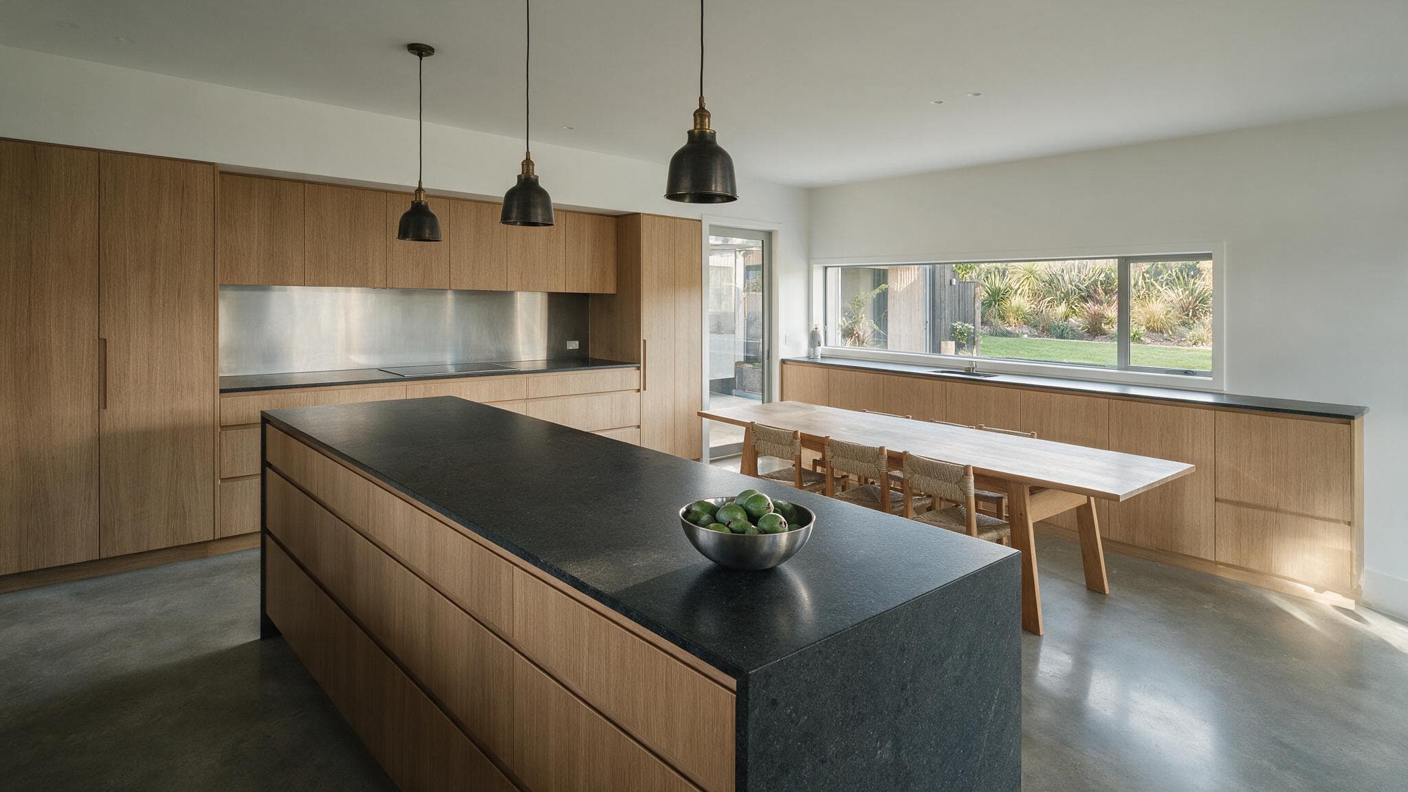 Interior render of a kitchen with full-height timber joinery, a dark stone island bench, three black pendant lights and a long dining table, with a window onto a garden.